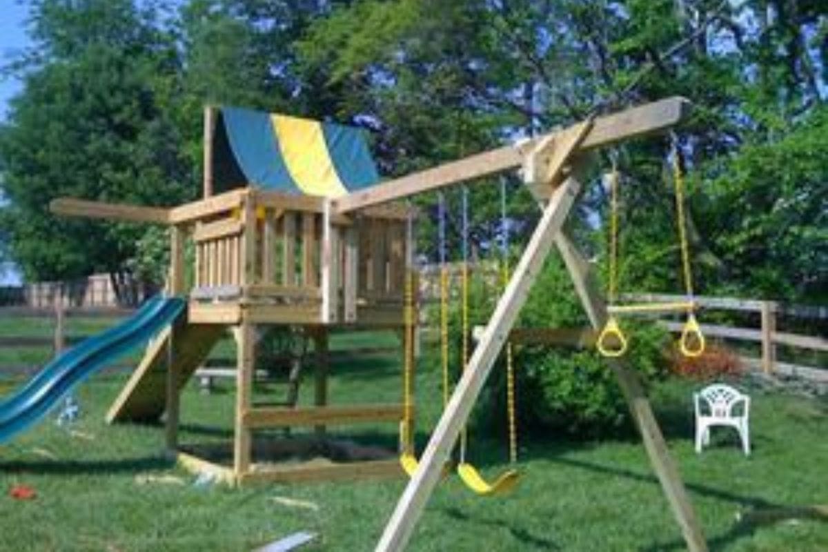 Wooden backyard playset with a slide, swings, climbing rings, and a platform with a fabric canopy-backyard play equipment worth investment-set on grass near a white plastic chair and trees.