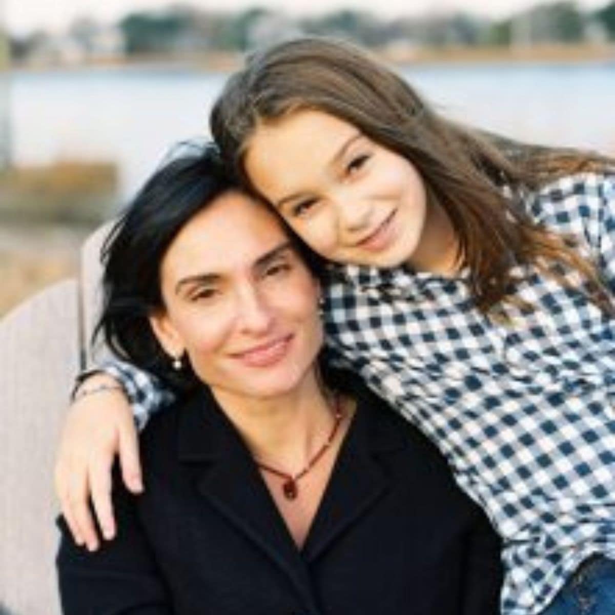 A woman and a young girl, Willa, sit closely together outdoors, both smiling at the camera. The girl rests her head on the woman’s shoulder as they enjoy a peaceful moment by the water in the background.