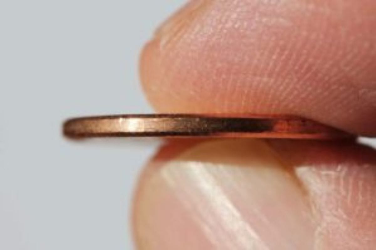 A close-up of a person's fingers holding the edge of a thin, copper-colored coin against a plain background, capturing a simple moment of saving.