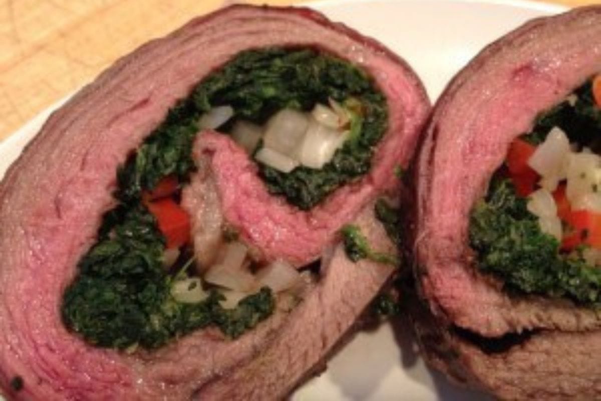 Spinach stuffed flank steak rolled with onions and red peppers, sliced and served on a white plate.