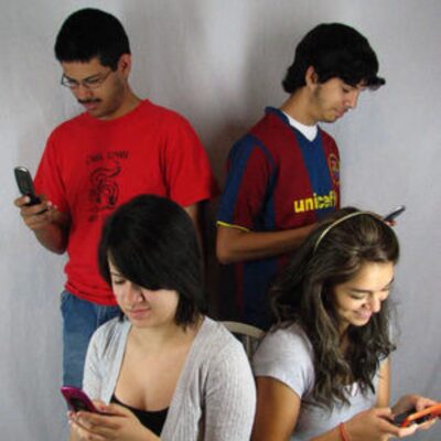 Four young people are standing and sitting, each looking down and using their smartphones, fully absorbed in technology against a plain background.