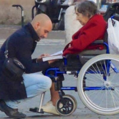 A man kneels beside an elderly woman in a wheelchair, holding a book and appearing to read or explain something to her—capturing one of life’s simple acts of kindness.