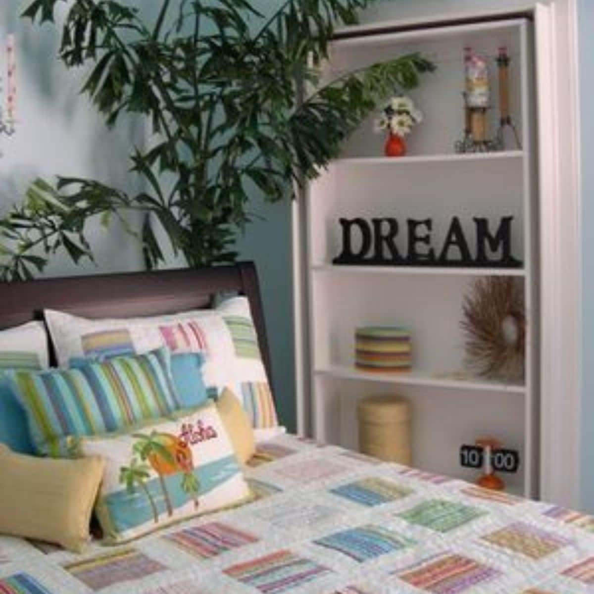 A neatly made bed with colorful striped pillows and quilt awaits guests, sitting next to a bookshelf displaying decorative items, including the word "DREAM" and a small clock showing 10:00.