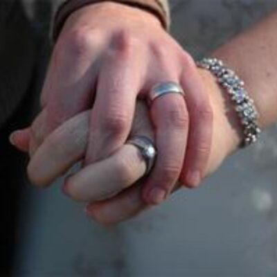 Two hands with silver rings are clasped together in a gentle gesture, symbolizing marriage; one hand also wears a silver bracelet.