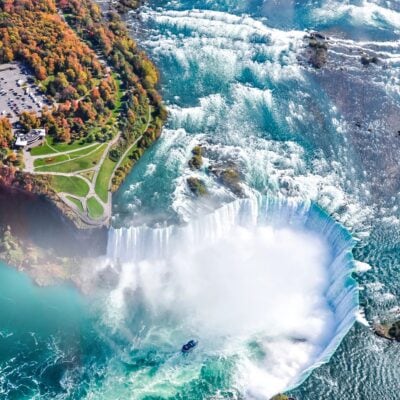 Aerial view of Niagara Falls with cascading water, mist rising, a tour boat near the base, and autumn trees and a parking lot visible on the shore at Niagara Falls.