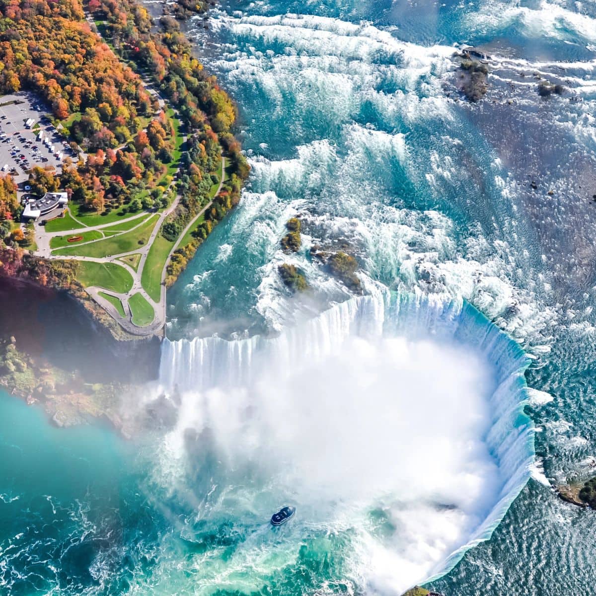 Aerial view of Niagara Falls with cascading water, mist rising, a tour boat near the base, and autumn trees and a parking lot visible on the shore at Niagara Falls.