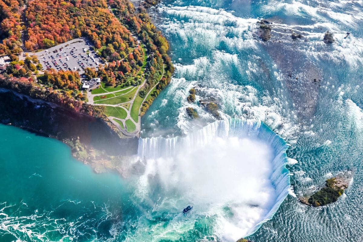 Aerial view of Niagara Falls with a tour boat near the base, vibrant autumn foliage on the left, and niagara falls parking lot and pathways visible.