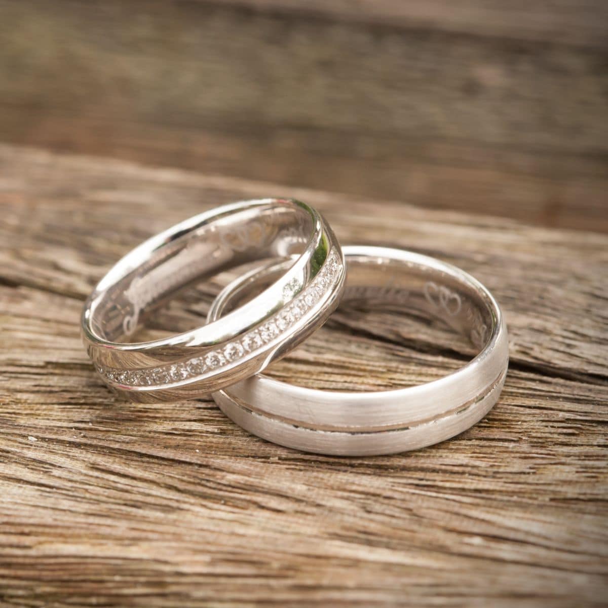 Two silver wedding rings rest on a wooden surface, with one wedding band featuring small embedded stones and the other remaining plain.