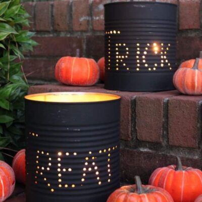 Two black tin cans with "TRICK" and "TREAT" spelled out in holes, lit from inside, serve as charming DIY Halloween decor on brick steps, surrounded by small decorative pumpkins.
