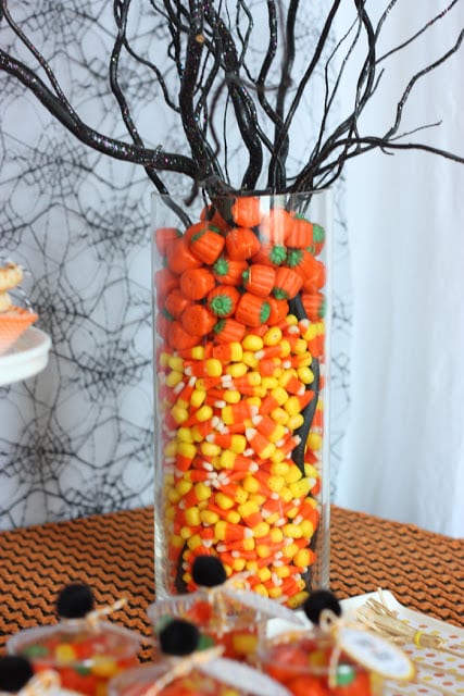 candy corn and marshmallow pumpkins in a glass vase with branches sticking out of it