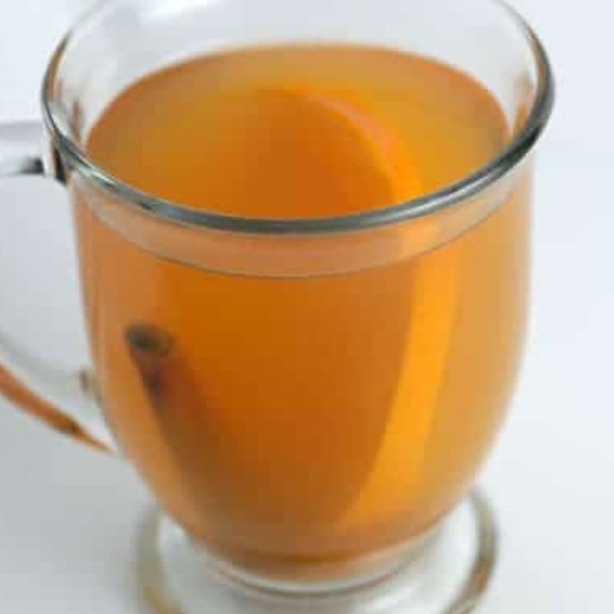A clear glass mug filled with light brown tea, reminiscent of apple cider, containing a slice of orange and a cinnamon stick.