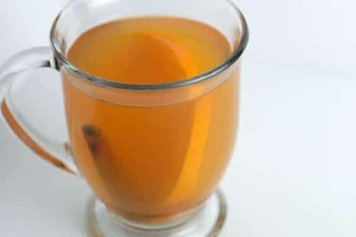 A clear glass mug filled with light brown tea, reminiscent of apple cider, containing a lemon wedge and a cinnamon stick, on a white surface.