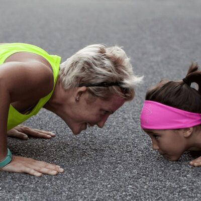 An adult and a child wearing athletic clothes face each other while doing push-ups on an outdoor surface, showing what it means to be a great sports mom.