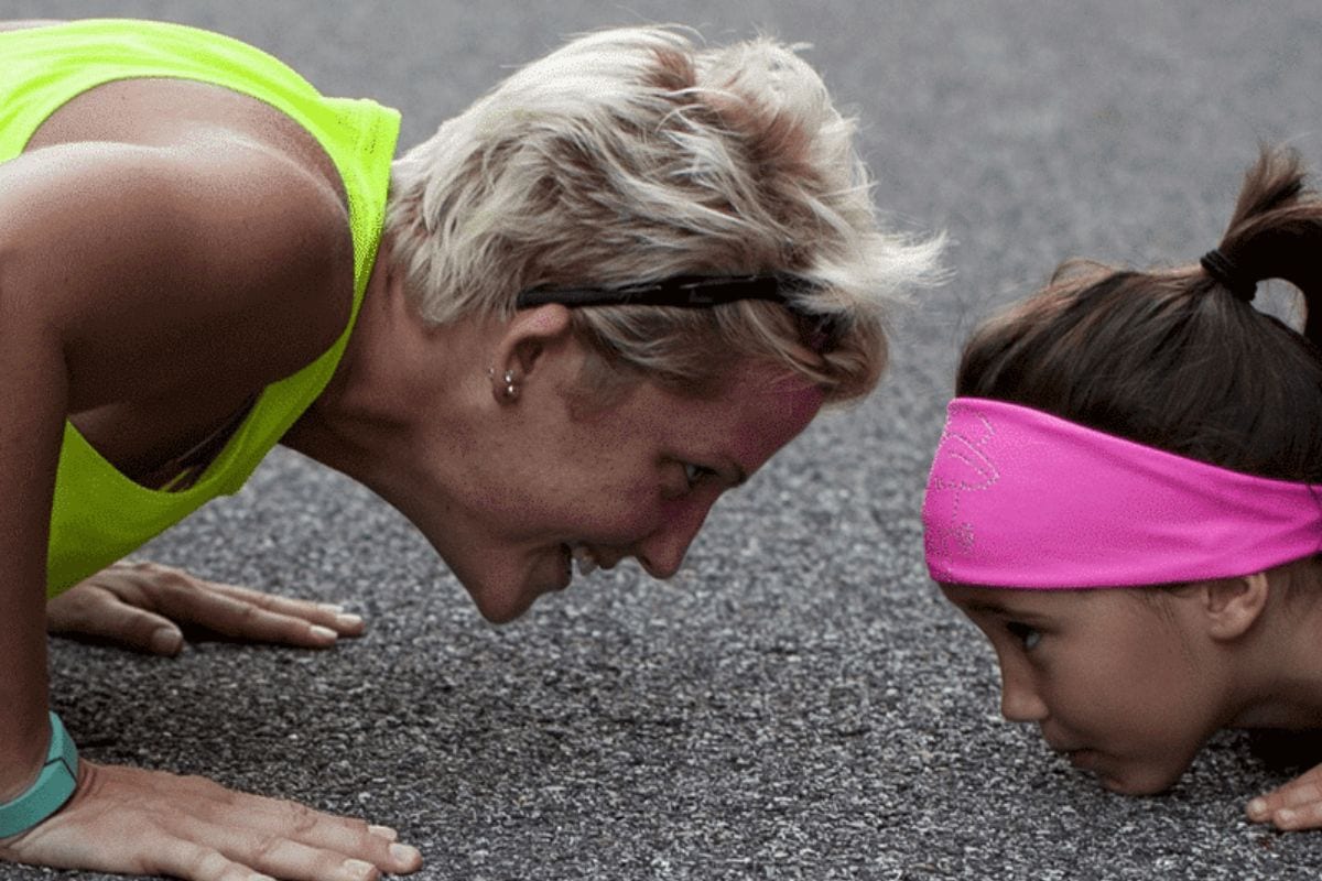 An adult and a child face each other while doing push-ups on a paved surface; both are focused and wearing athletic clothing-a true great sports mom moment in action.
