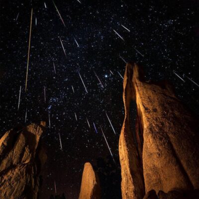 Tall rock formations stand beneath a night sky illuminated by numerous meteor streaks during a spectacular meteor shower.