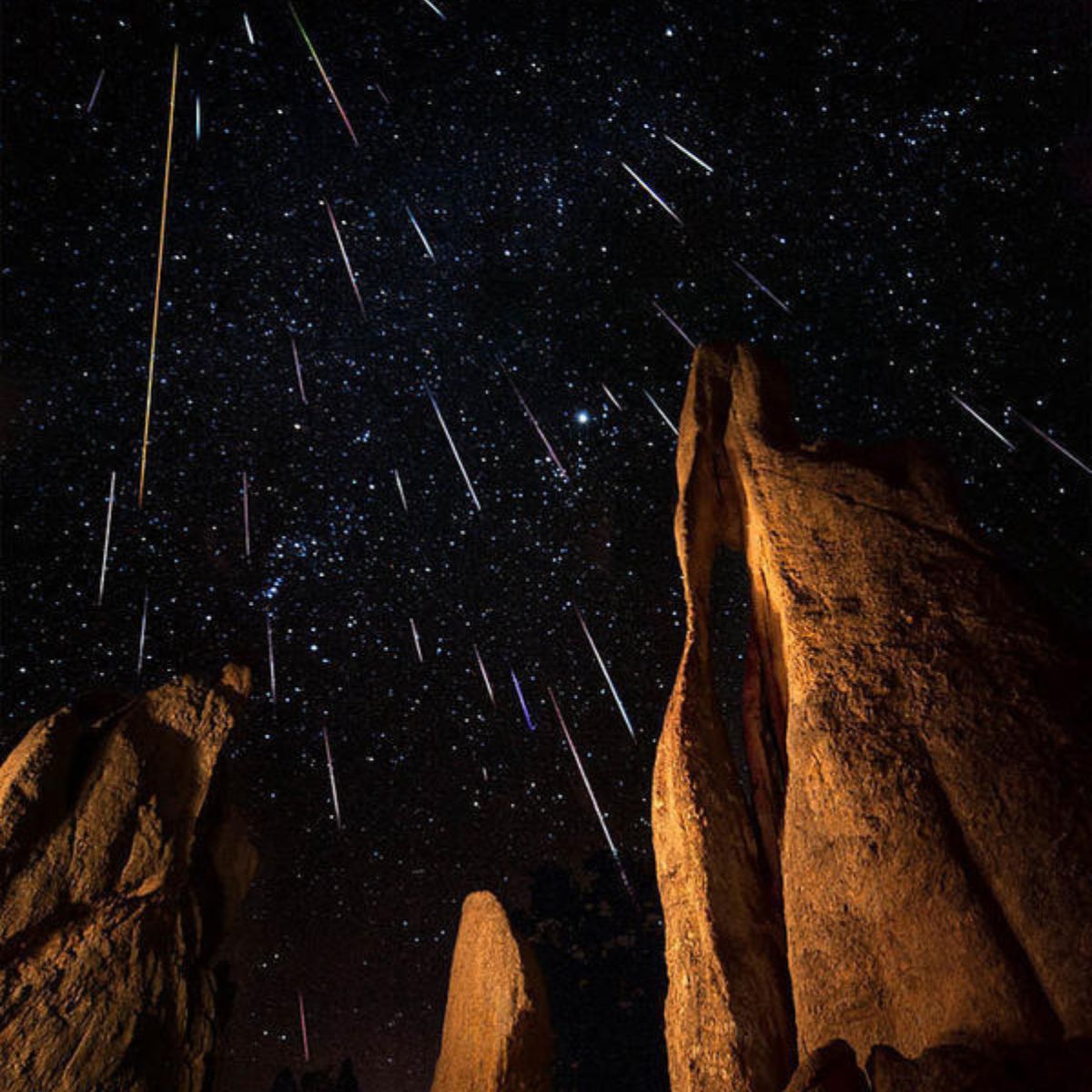 Tall rock formations stand beneath a night sky illuminated by numerous meteor streaks during a spectacular meteor shower.