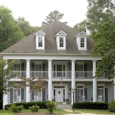 A large, two-story house with gray siding, white pillars, dormer windows, and a covered front porch with rocking chairs and an American flag—perfect for peace of mind with homeowners insurance.
