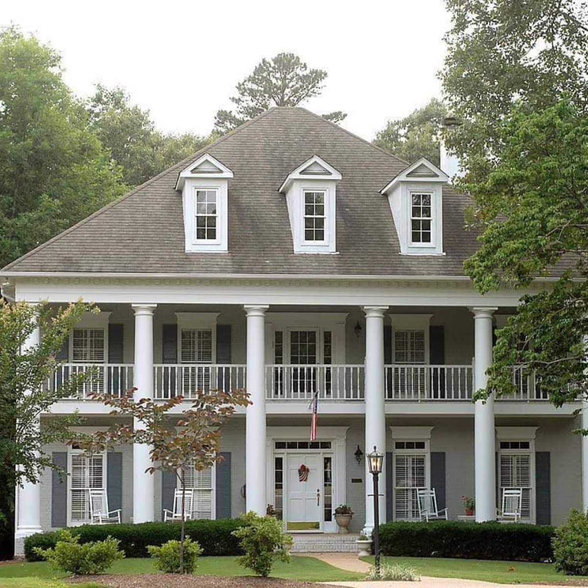 A large, two-story house with gray siding, white pillars, dormer windows, and a covered front porch with rocking chairs and an American flag—perfect for peace of mind with homeowners insurance.