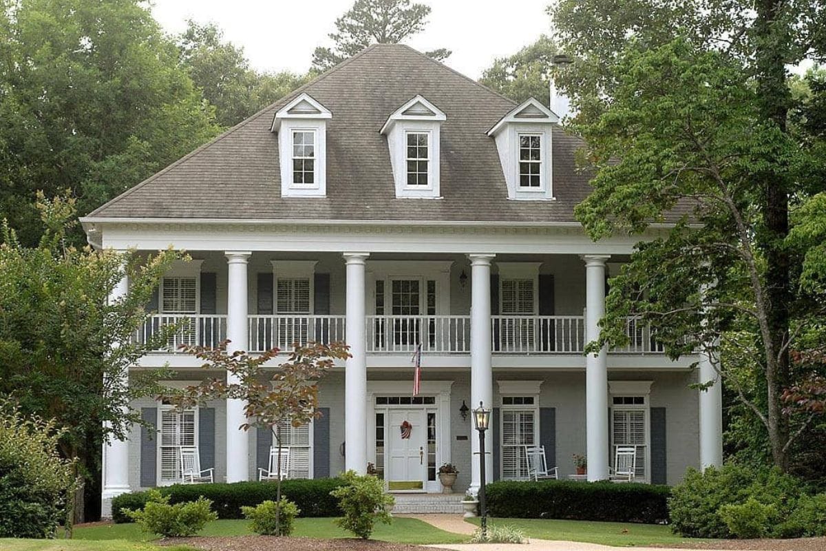 A two-story house with white columns, a wide front porch, dormer windows, and rocking chairs, surrounded by trees and greenery-a perfect haven protected by reliable homeowners insurance.