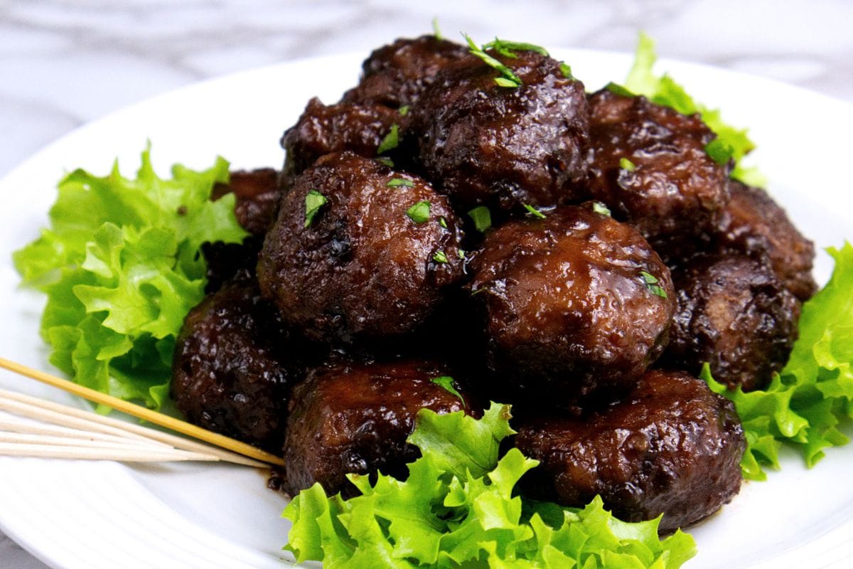 These easy sweet & sour meatballs are a party favorite! They're super easy to make and guests always ask for the recipe. #appetizers #fingerfoods #meatballs A plate of tender glazed meatballs garnished with chopped herbs, served on a bed of fresh green lettuce leaves.