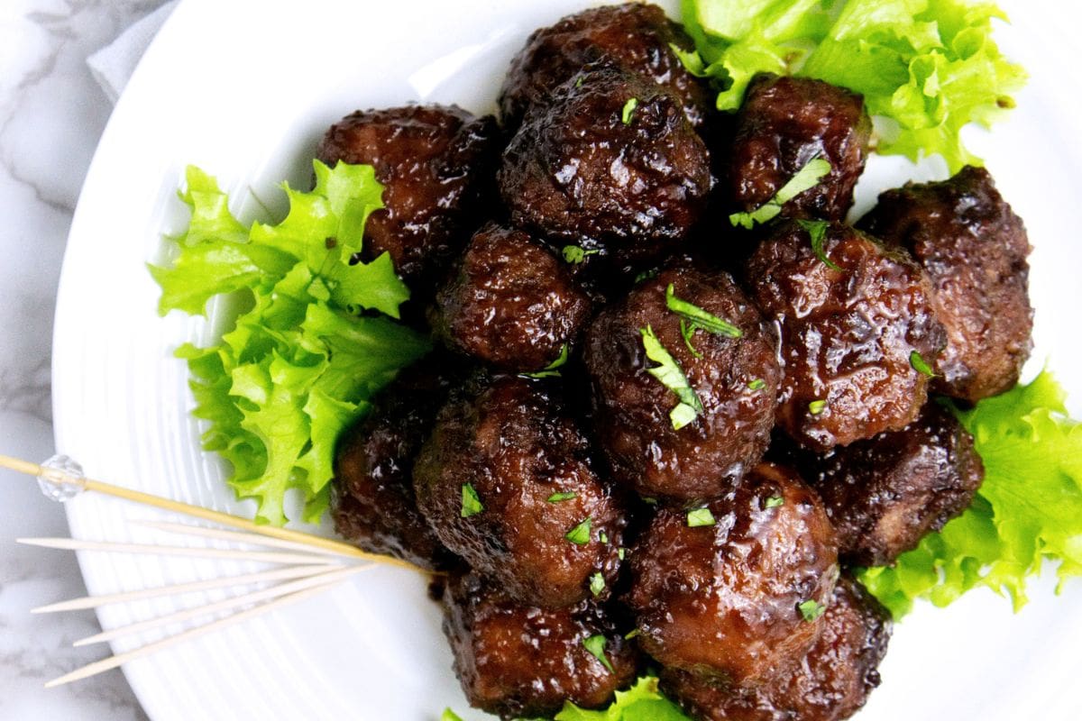 These easy sweet & sour meatballs are a party favorite! They're super easy to make and guests always ask for the recipe. #appetizers #fingerfoods #meatballs A plate of savory glazed meatballs garnished with chopped herbs and leafy lettuce, served with cocktail toothpicks on the side.