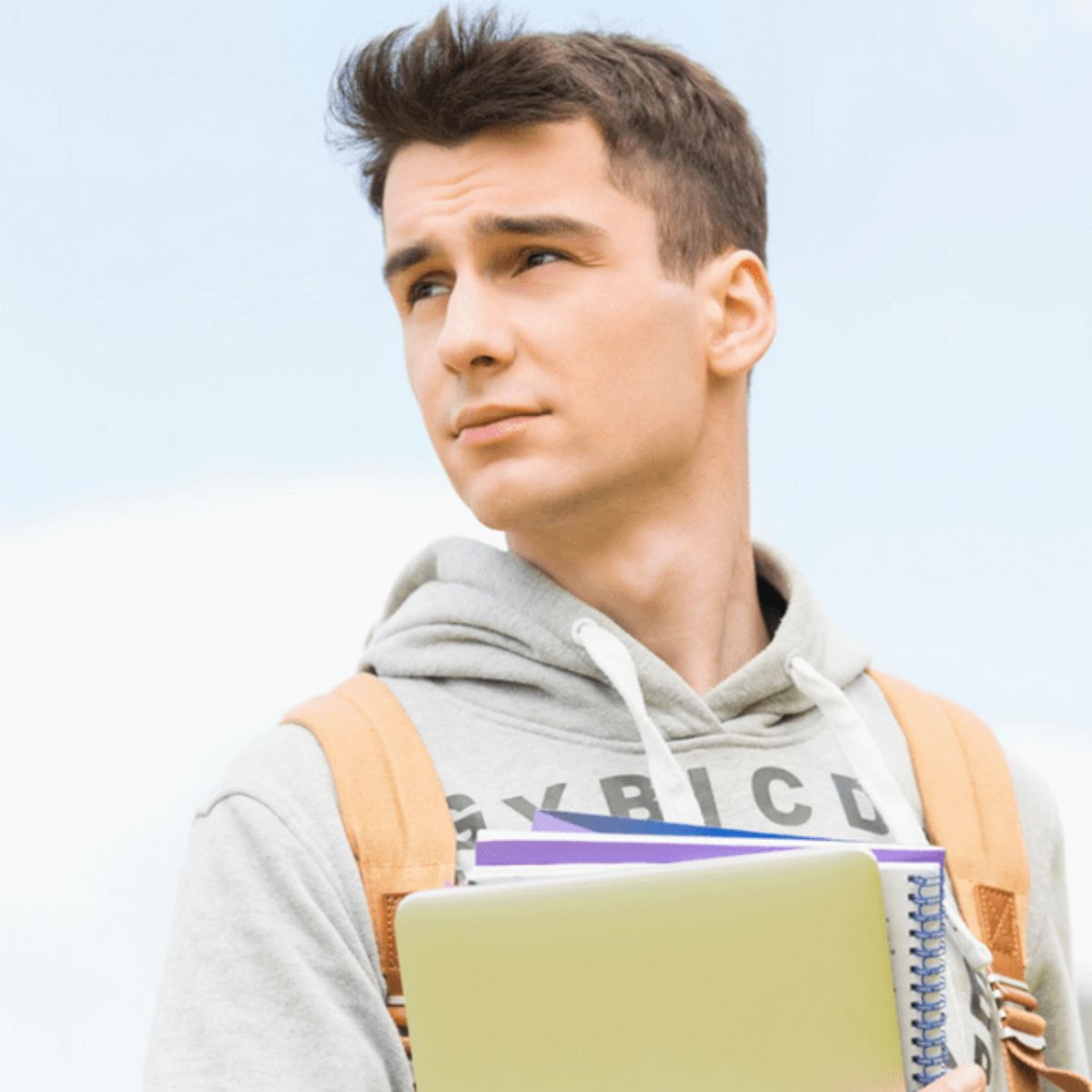 Young man wearing a gray hoodie and backpack holds a notebook, spiral-bound folders, and what appears to be a college admission résumé, looking off to the side against a light background.