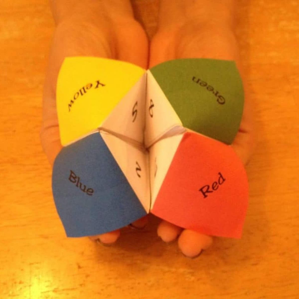 Two hands hold a printable fortune teller with four flaps labeled yellow, green, blue, and red, displaying numbers and colors on each section.
