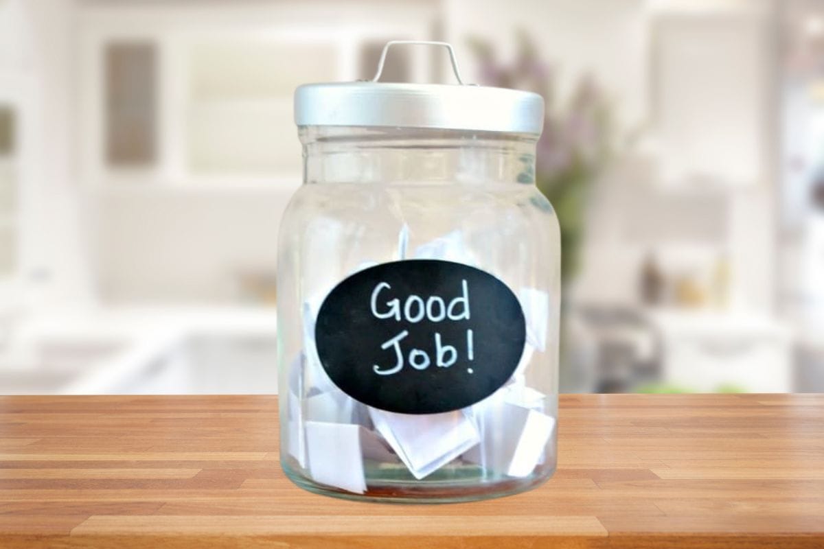 A glass jar labeled "Good Job!" filled with folded paper slips, like kid coupons, sits on a wooden countertop.