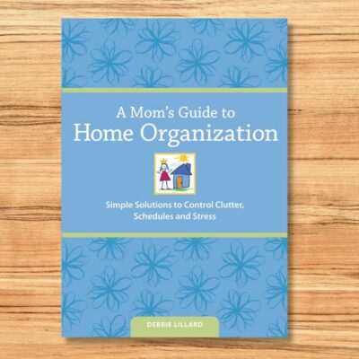 The cover of "A Mom’s Guide to Home Organization" by Debbie Lillard rests on a wooden surface, adorned with blue floral designs and a simple house illustration, perfectly capturing the essence of a mom's guide to home organization.