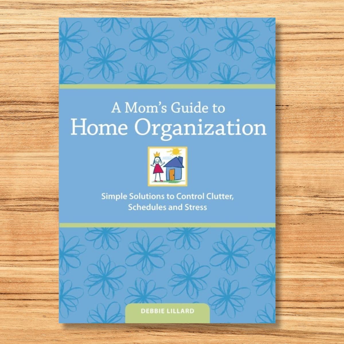 The cover of "A Mom’s Guide to Home Organization" by Debbie Lillard rests on a wooden surface, adorned with blue floral designs and a simple house illustration, perfectly capturing the essence of a mom's guide to home organization.