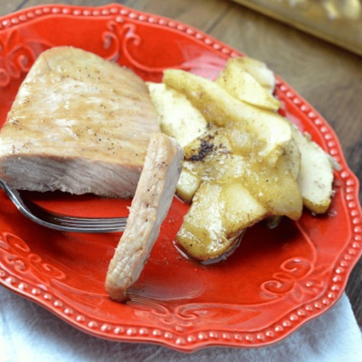 A piece of baked pork chops and apples, with a slice cut, is served on a red plate alongside tender cooked apple slices.