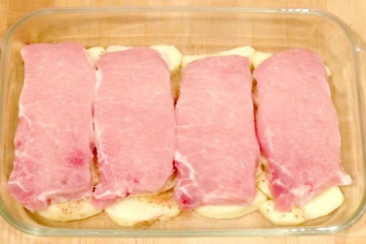 Four raw pork chops arranged in a glass baking dish on top of sliced potatoes, perfect for making baked pork chops and apples.