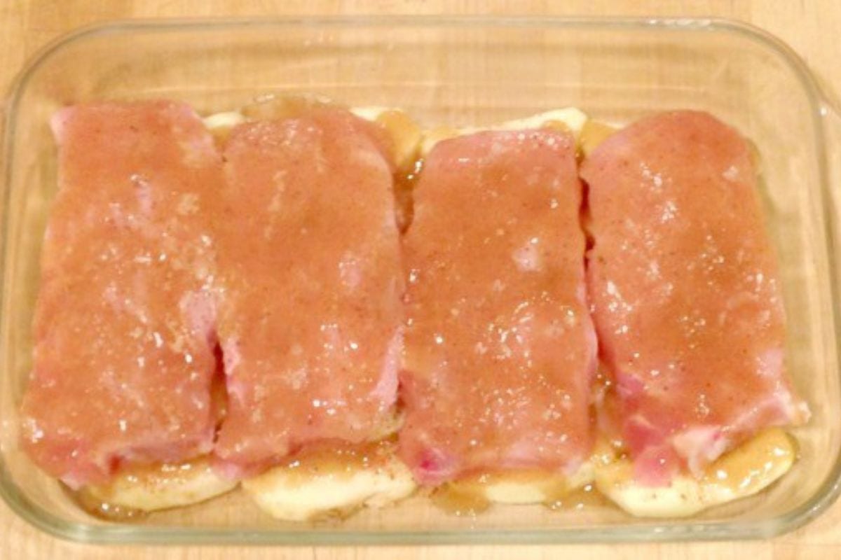 Four raw pork chops coated with a brown sauce are arranged in a glass baking dish on a wooden surface.