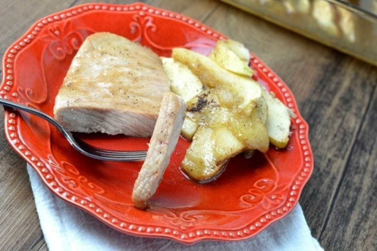 A red plate with baked pork chops and apples, alongside a fork, sits on a wooden surface.