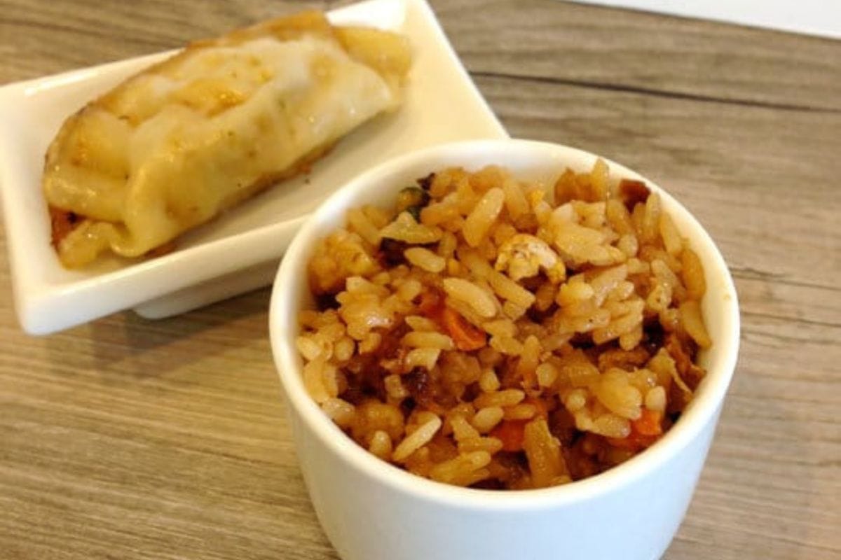 A bowl of fried rice with vegetables and bits of egg sits next to a single dumpling in a small rectangular dish on a wooden surface-perfect for those seeking festive Chinese New Year recipes.