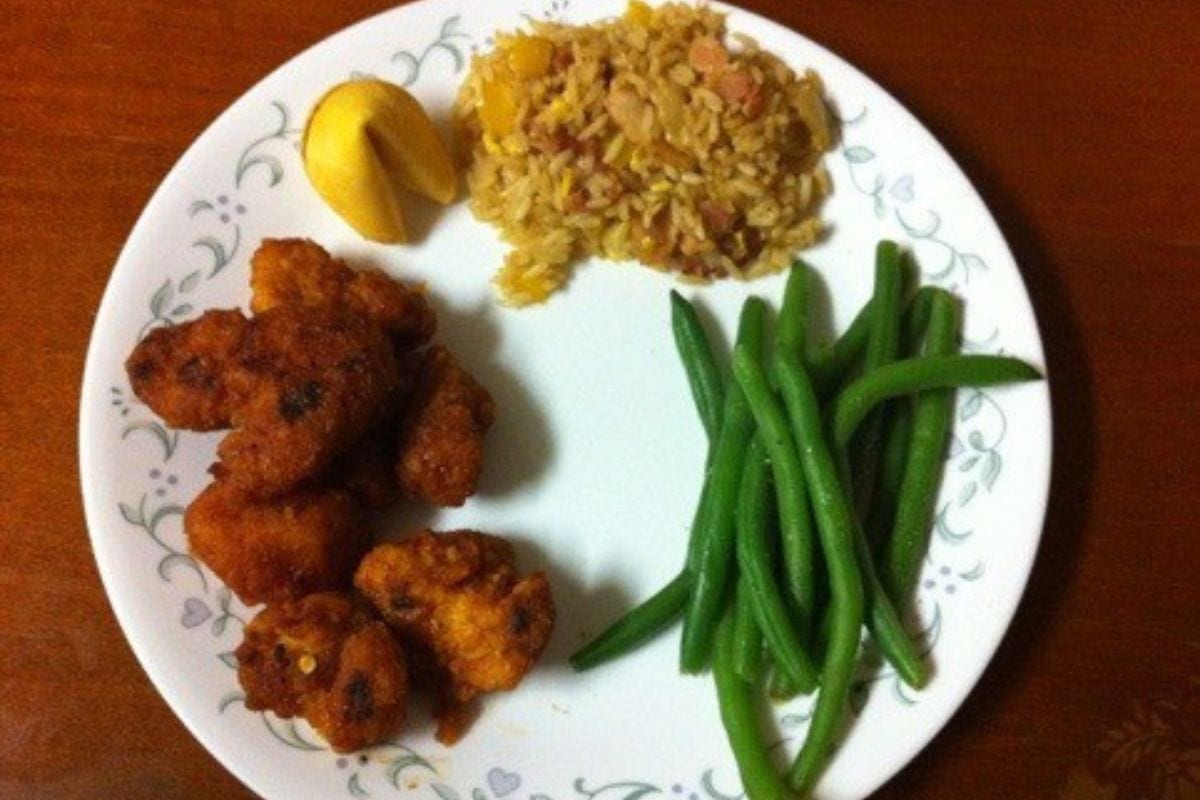 A plate with fried chicken pieces, green beans, a serving of rice with mixed ingredients, and a lemon wedge on a white plate with a floral border-perfect for celebrating with Chinese New Year recipes.