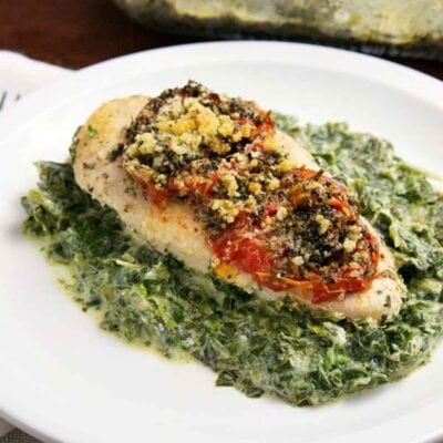 A chicken florentine bake features a baked chicken breast topped with tomatoes and herbs, served on a bed of creamy spinach on a white plate.