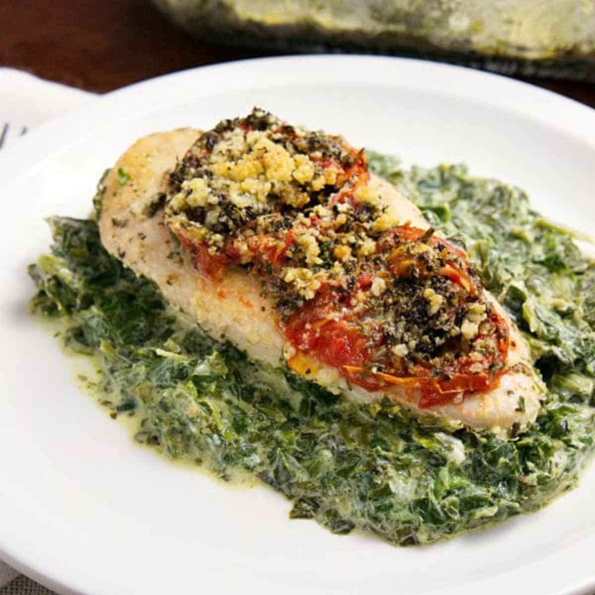 A chicken florentine bake features a baked chicken breast topped with tomatoes and herbs, served on a bed of creamy spinach on a white plate.