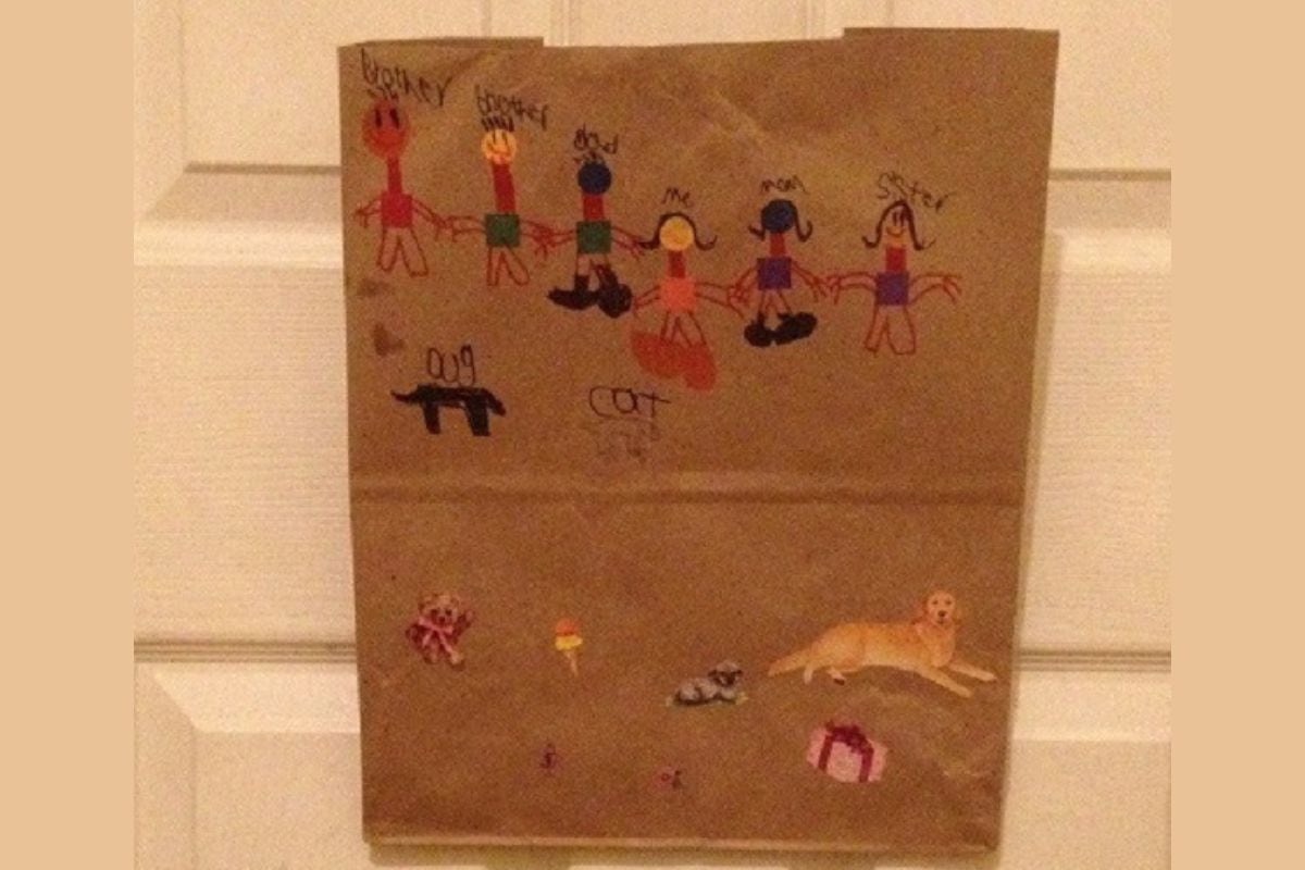 A child's drawing on brown paper shows five people labeled as family members, a black dog, a cat, and animal stickers including a dog, family mailboxes, and a wrapped gift at the bottom.