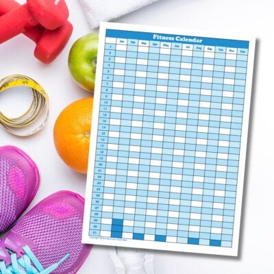 A blank fitness calendar is placed on a white surface next to pink dumbbells, measuring tape, green apple, orange, pink running shoes, and a towel—everything you need to plan and improve your fitness.
