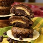 A stack of peanut butter cup chocolate cookies with one cut in half, showing the rich filling. The cookies are placed on a patterned cloth with some chocolate chips scattered around.