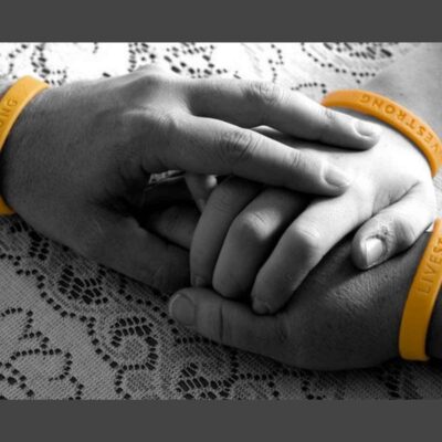 Two hands wearing yellow "LIVESTRONG" wristbands, symbolizing strength in the fight against cancer, gently hold the smaller hand of a child on a lace tablecloth.