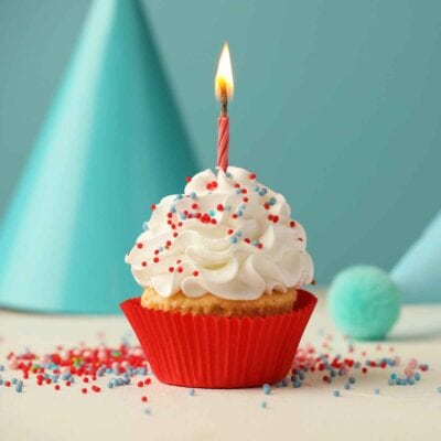 A cupcake with white frosting, red and blue sprinkles, and a lit birthday candle in a red wrapper features neapolitan designs. Blue party hats and decorations are in the background.