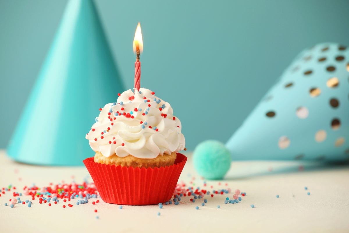 A cupcake with white frosting, red and blue sprinkles, and a lit candle sits in a red wrapper.