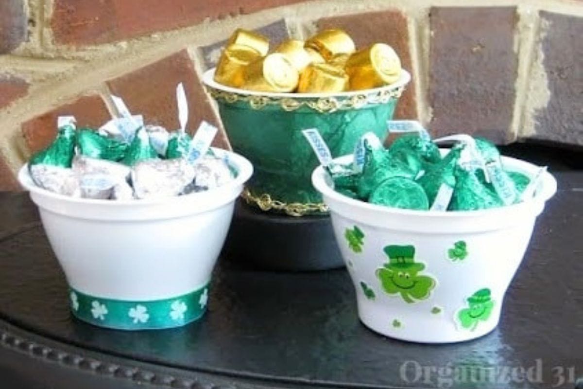 Three decorated cups filled with assorted chocolates, candies, and creme de menthe brownies are artfully arranged on a dark surface in front of a brick wall.