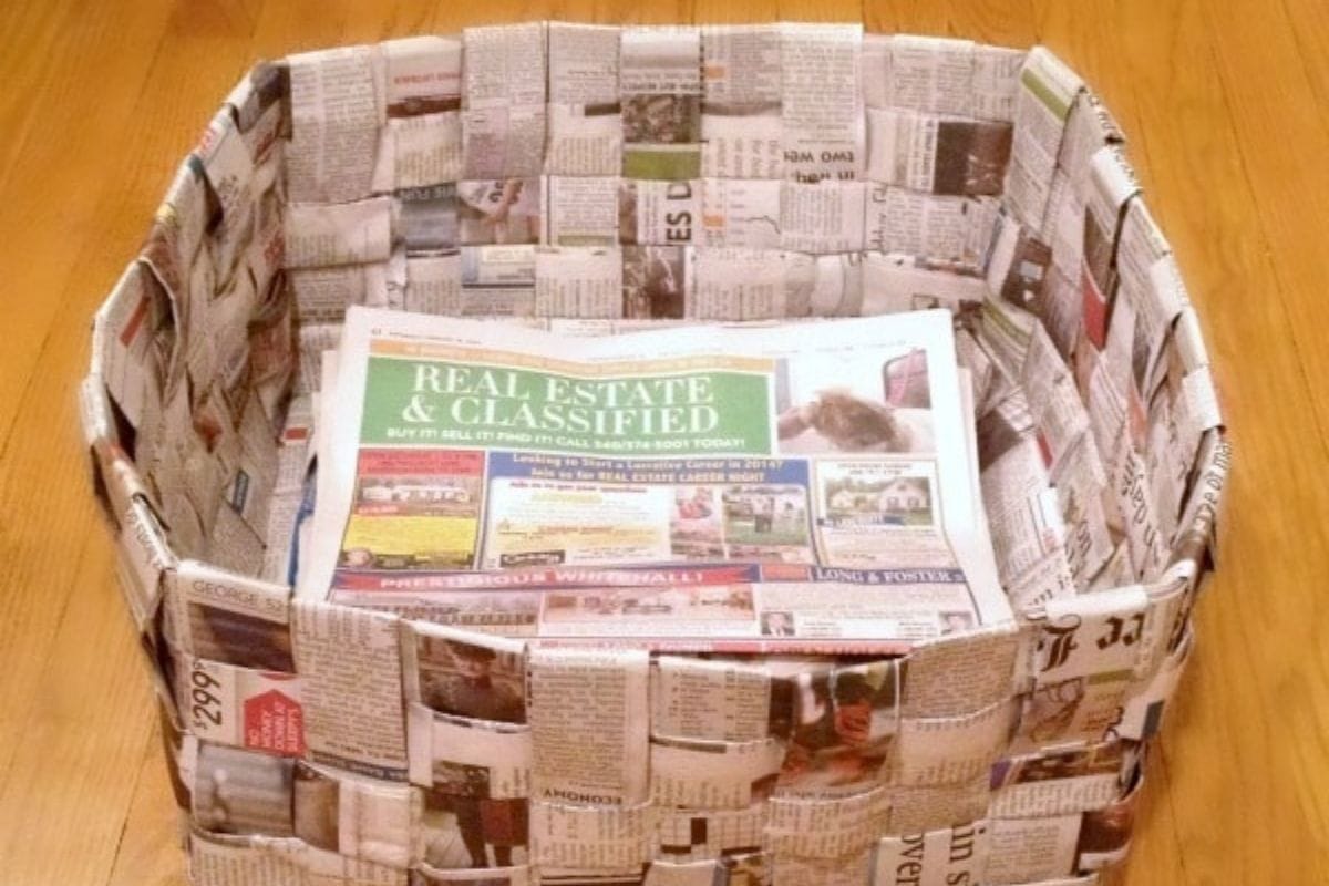 A rectangular newspaper basket woven from strips of paper sits on a wooden floor, with a folded newspaper lying inside the basket.