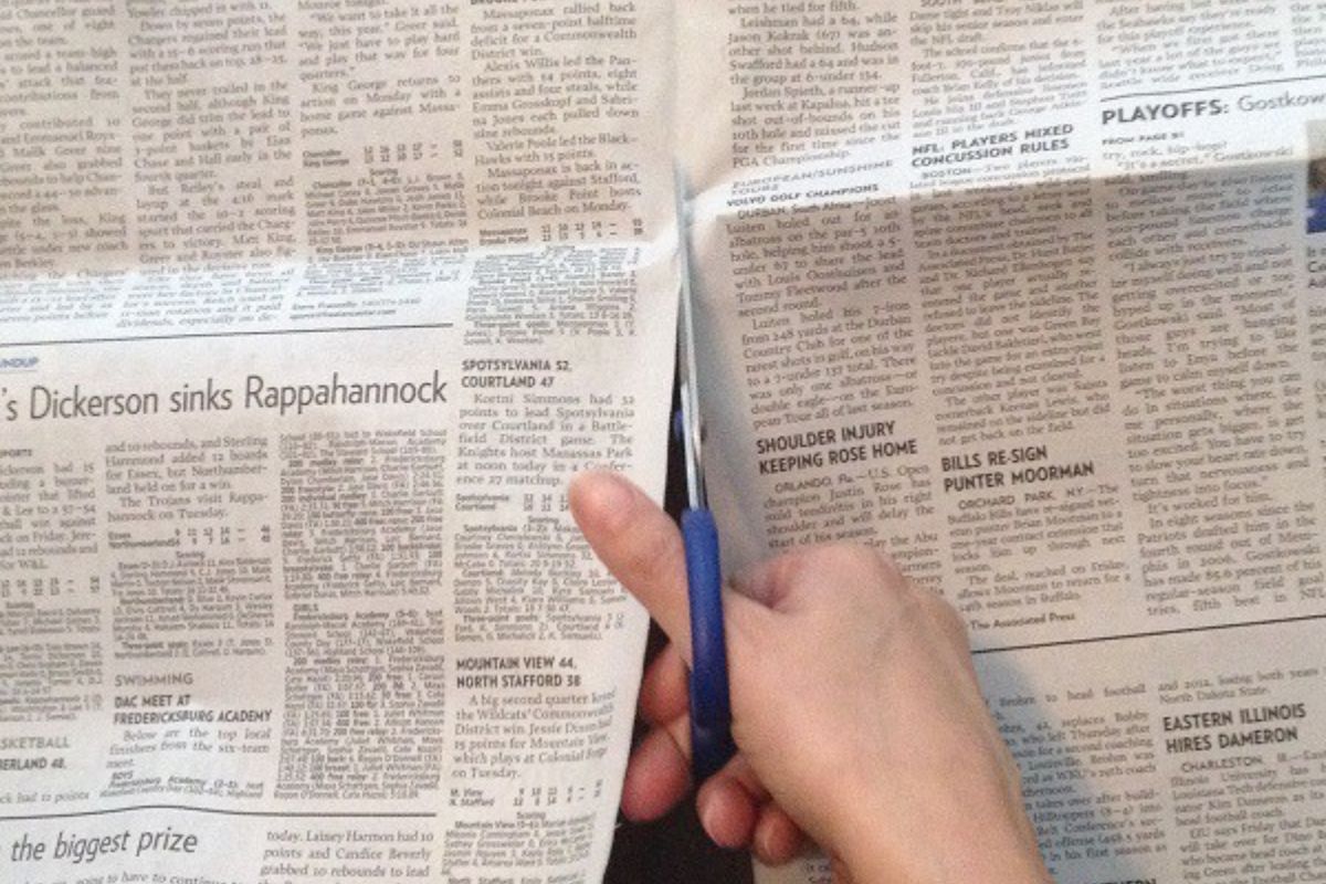 A person cuts a newspaper in half using blue-handled scissors, with various sports articles visible on the page-perhaps preparing materials for a creative newspaper basket.