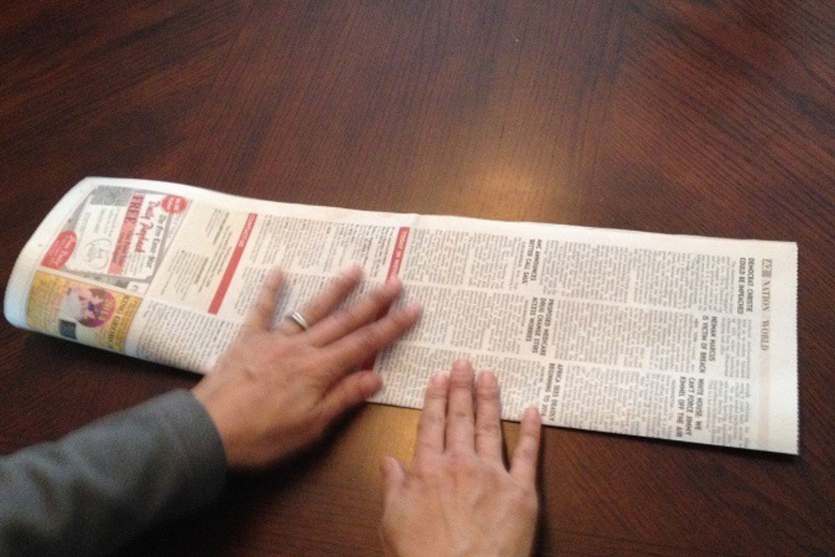 Two hands are folding a newspaper lengthwise on a wooden table, preparing strips to weave into a newspaper basket.