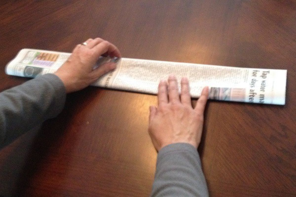 A person folding a newspaper lengthwise on a wooden table, preparing strips to weave into a newspaper basket.