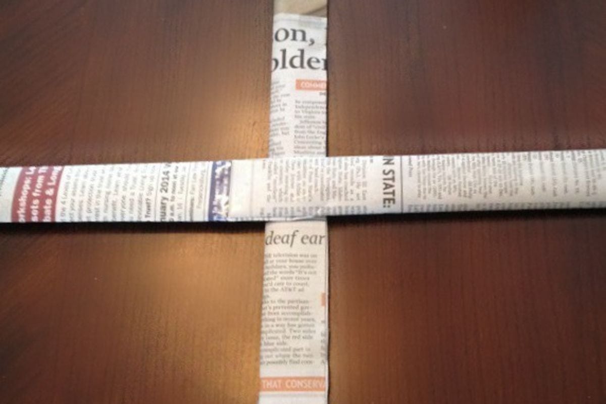 Two folded newspapers are arranged perpendicularly on a wooden surface, forming a cross shape reminiscent of starting a newspaper basket.